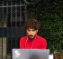Man with curly hair and headphones using a laptop in front of a gated window and leafy green hedge. He's wearing a red shirt and has a focused expression.