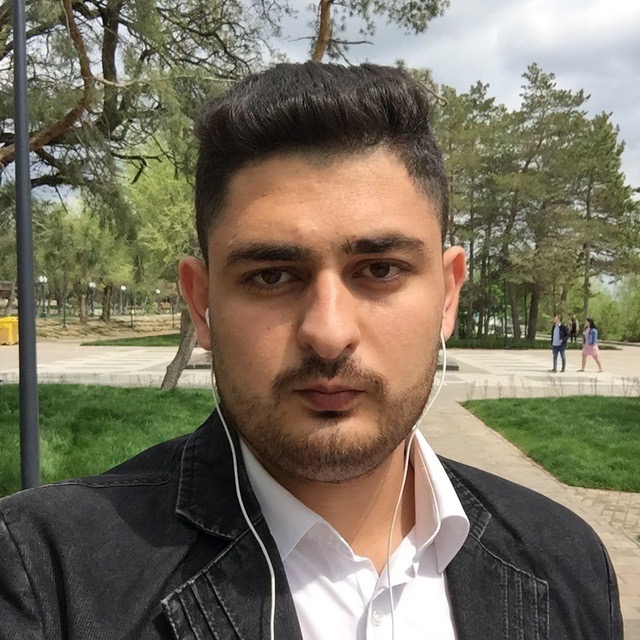 A young man wearing earphones, a black jacket, and a white shirt taking a selfie with a park in the background.