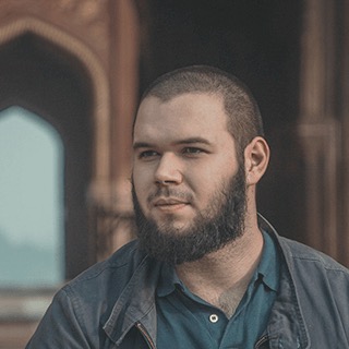 Bearded young man with short hair looking to the side with a historical building's arch in the background.