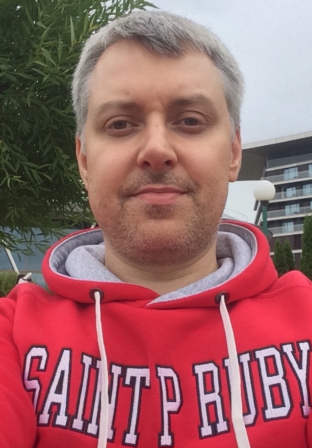 A selfie of a person with short grey hair wearing a red hoodie with the text "SAINT RUBY" in a casual outdoor setting with foliage and a building in the background.