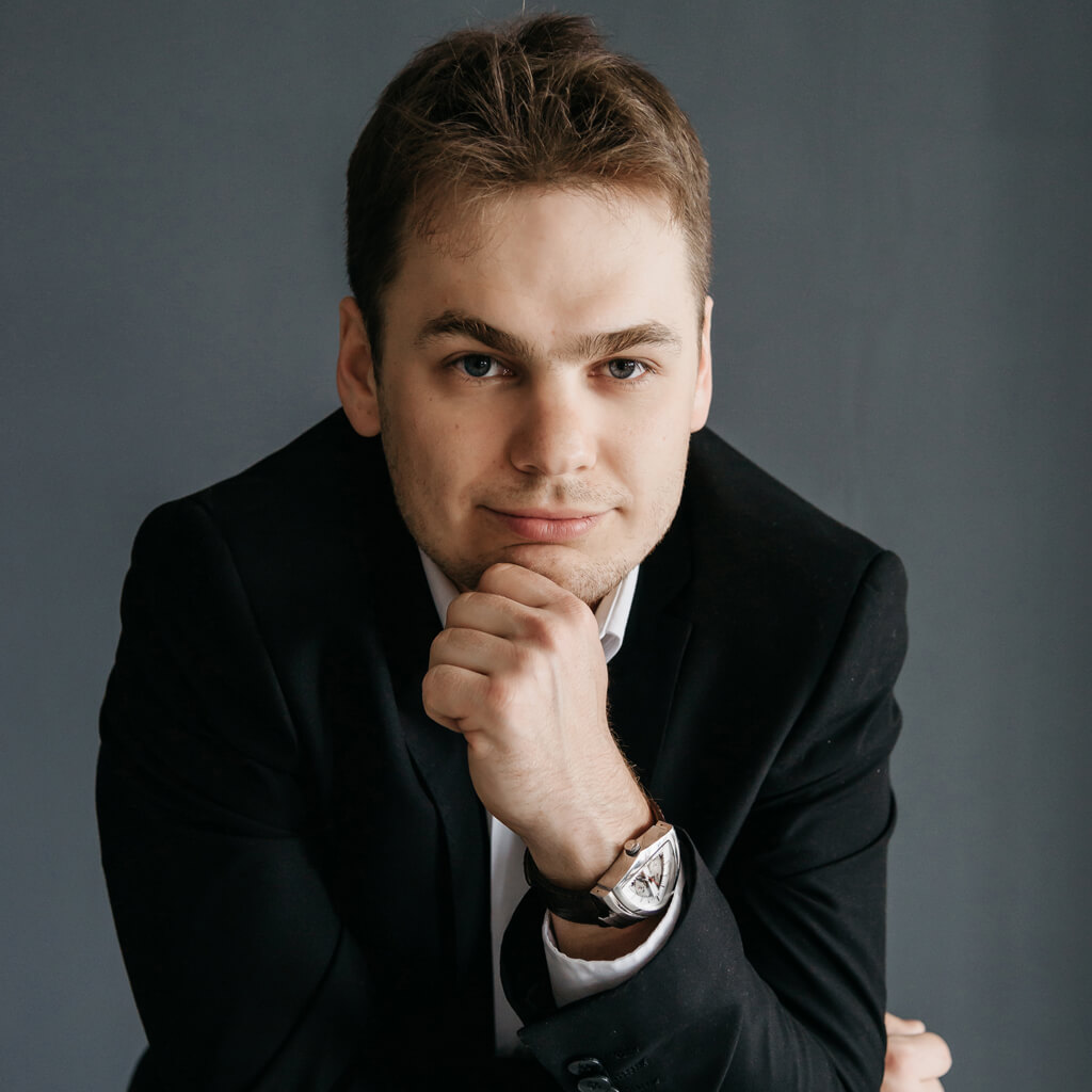 A portrait of a young man in a black suit resting his chin on his hand, wearing a wristwatch, and looking confidently at the camera against a grey background.