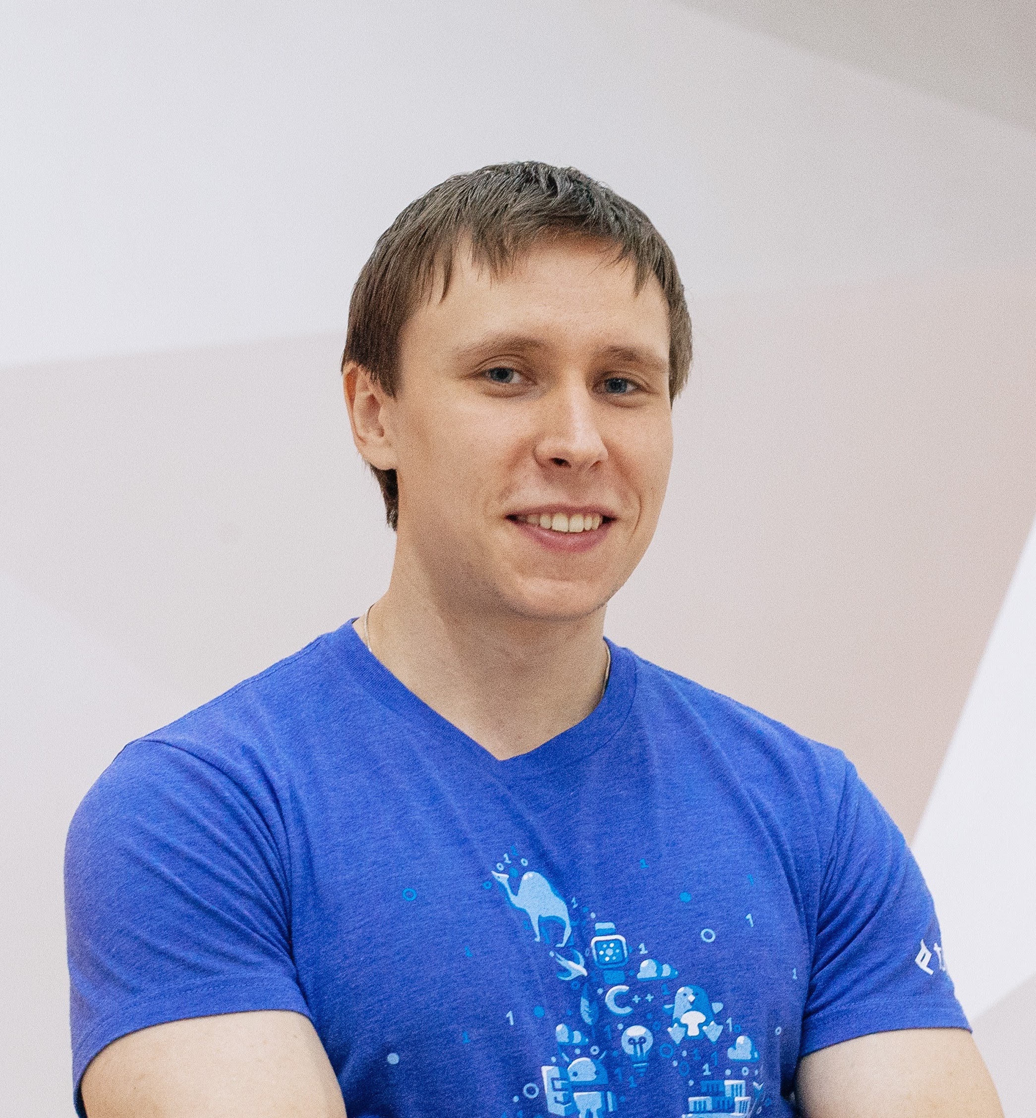 Smiling person in a blue T-shirt with a tech-themed graphic design, standing indoors with a neutral background.