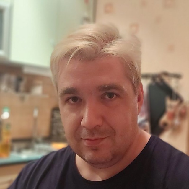 Portrait of a person with short blond hair in a blue top, indoors with a softly focused kitchen background.