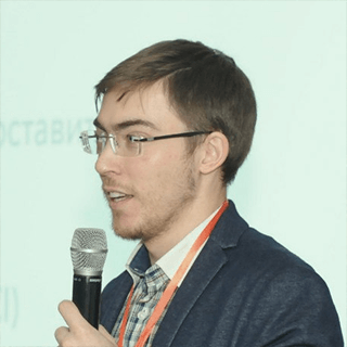 A man with glasses holding a microphone, presenting at an event, with Cyrillic text partially visible in the background.