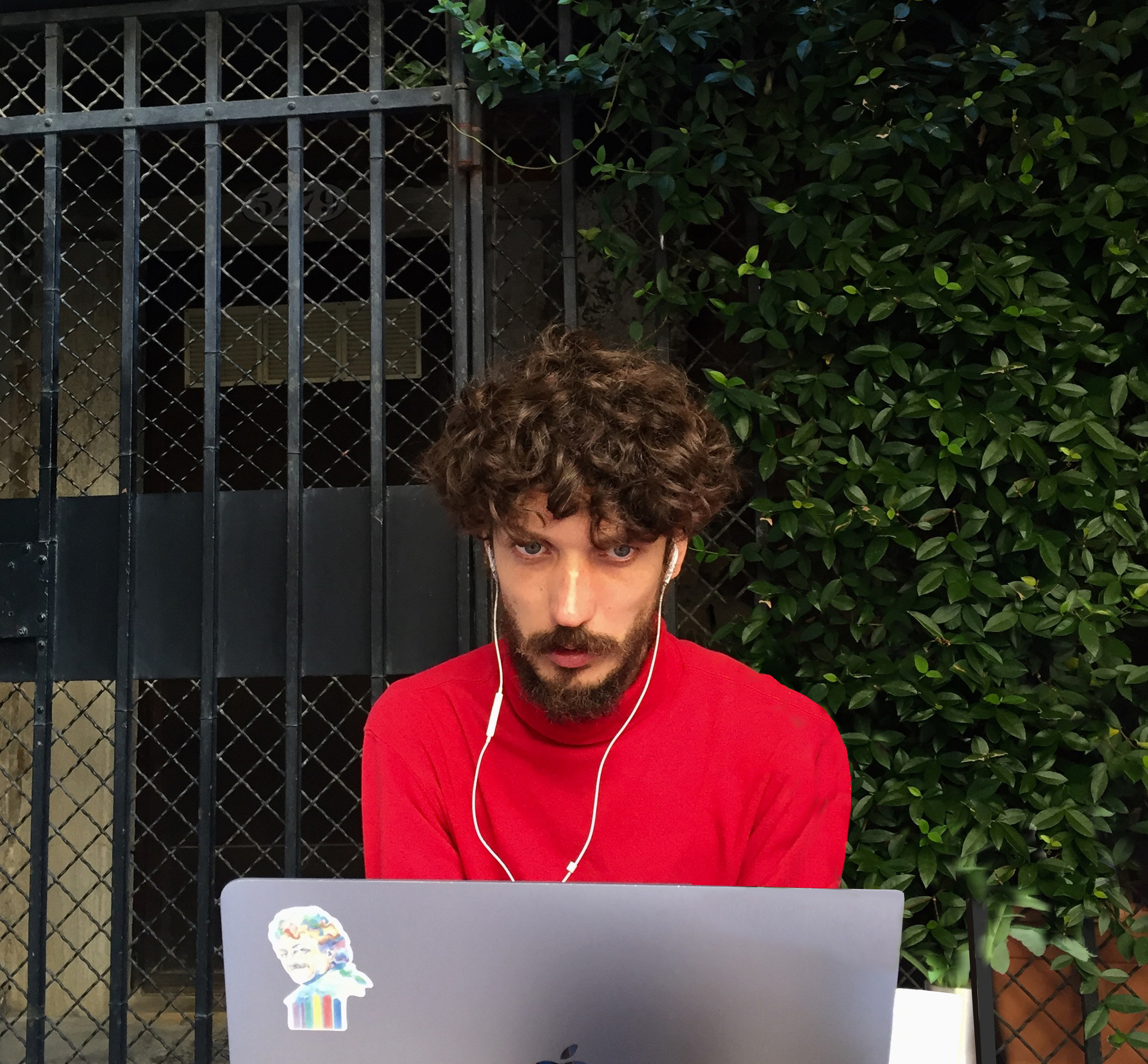 Man with curly hair and headphones using a laptop in front of a gated window and leafy green hedge. He's wearing a red shirt and has a focused expression.