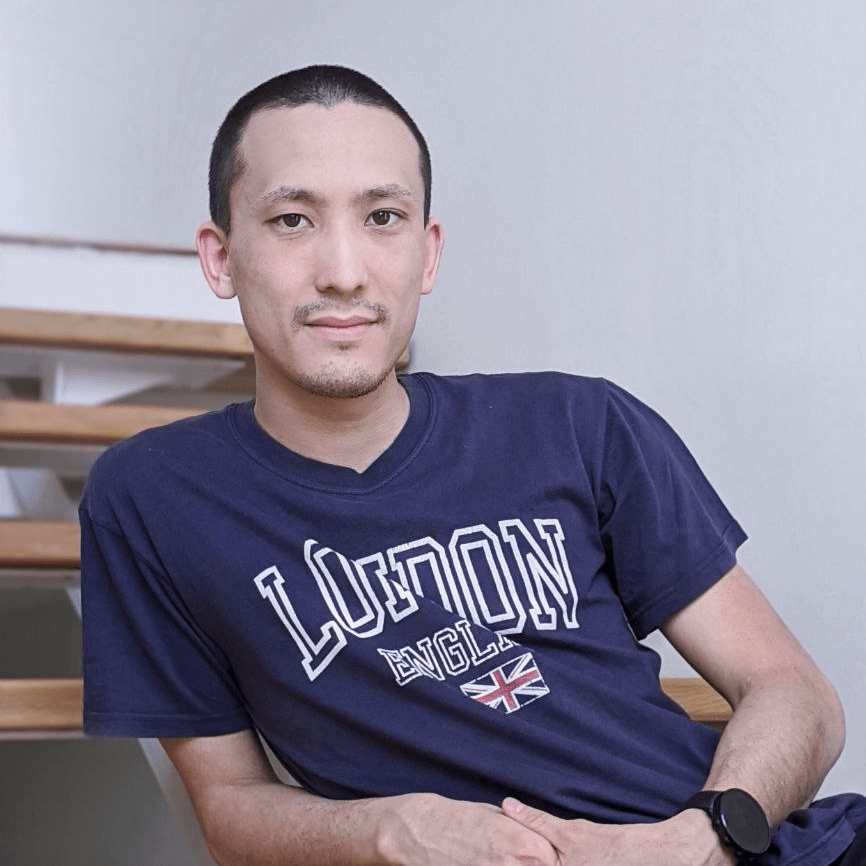 Man in a dark blue t-shirt with "London England" print, wearing a watch, sitting with crossed arms, with a stairway in the background.