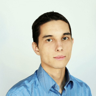 A young man wearing a blue shirt, with short hair, looking directly at the camera against a light grey background.