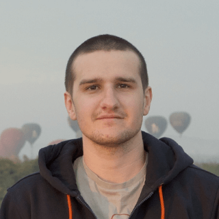 A portrait of a smiling man with short hair, wearing a hoodie, with blurry hot air balloons and a haze-covered landscape in the background.