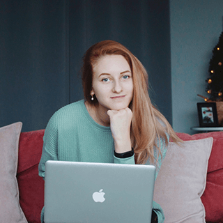 Woman in green sweater sitting on a couch with a laptop on her knees, resting chin on hand, with a decorated Christmas tree in the background.