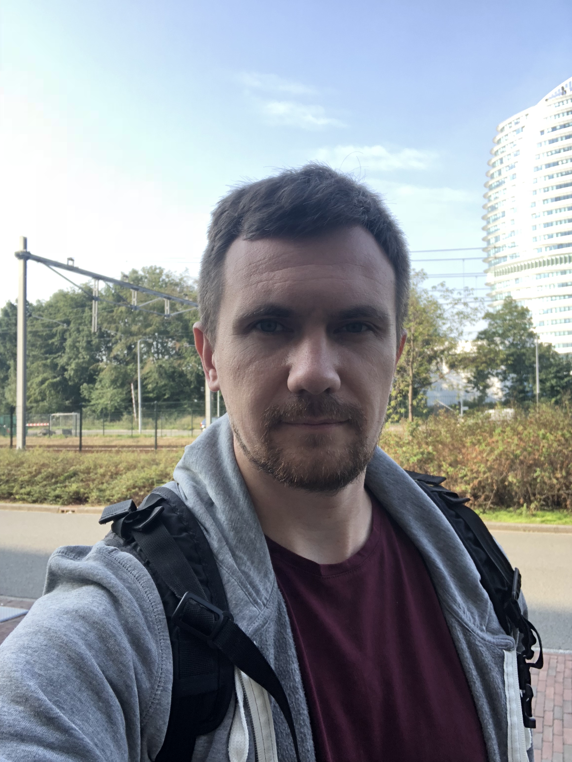 Selfie of a person outdoors with a grey hoodie and backpack, with buildings and greenery in the background.