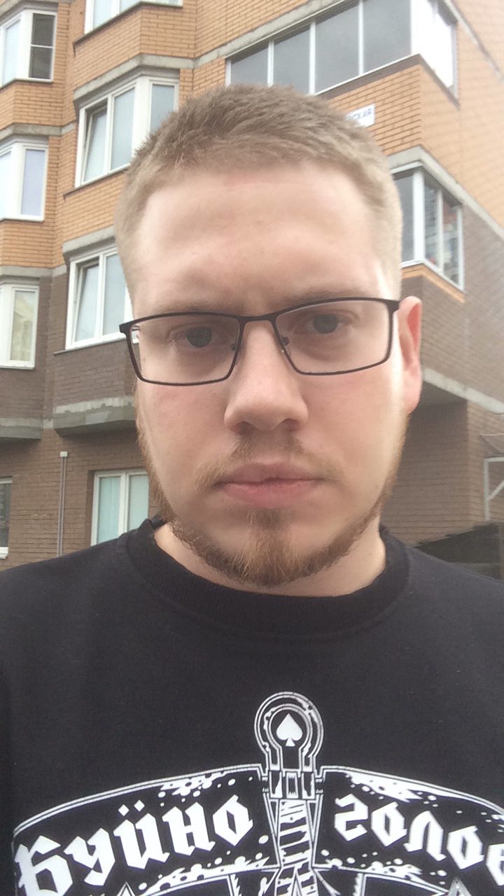 A close-up selfie of a young man with glasses, a short beard, and short hair, standing in front of a brick apartment building, wearing a black t-shirt with white text and graphics.