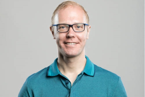 Smiling person with glasses wearing a teal polo shirt against a gray background.