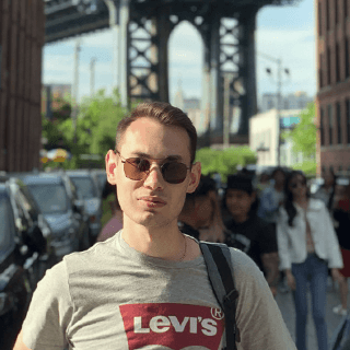 Man wearing sunglasses and a Levi's t-shirt standing in front of a city street with people and cars under a bridge structure on a sunny day.