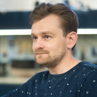 A man with a short beard and mustache, wearing a blue shirt with small patterns, is looking to the side with a slight smile, with an out-of-focus background suggesting an indoor setting.