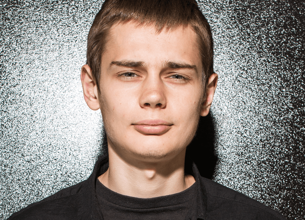 Close-up of a young male with a neutral expression against a speckled black and white background.