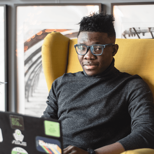 A young man wearing glasses and a black turtleneck sitting in a yellow armchair with a laptop covered in stickers, possibly working or studying.