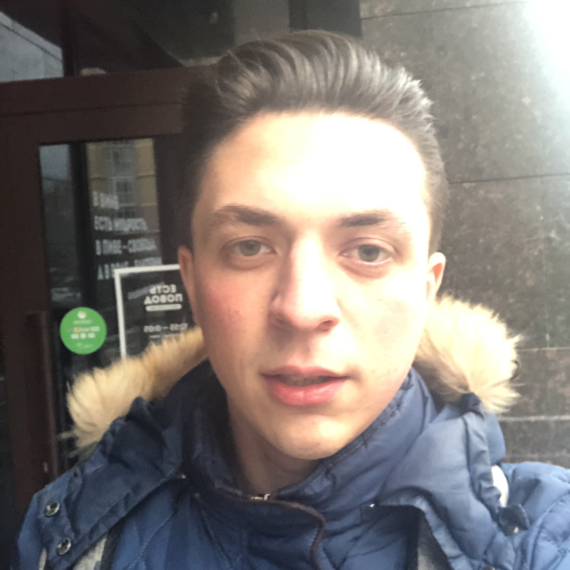 A close-up selfie of a young person with short hair wearing a blue winter jacket with a fur-lined hood, standing outside a building.