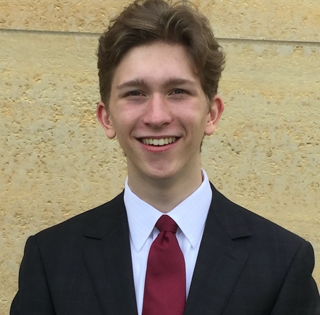 Young man with a cheerful smile wearing a black suit and red tie standing before a beige wall.