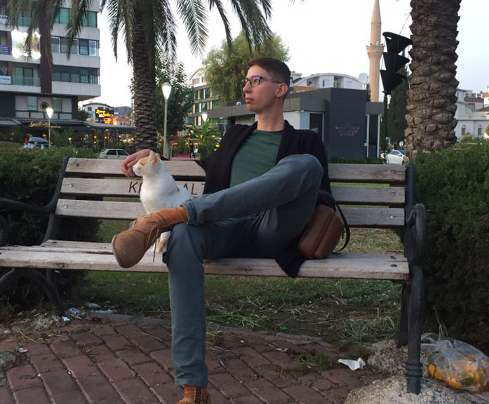 Man sitting on a park bench with a relaxed posture, petting a white cat, with trees, a mosque minaret, and city buildings in the background at dusk.