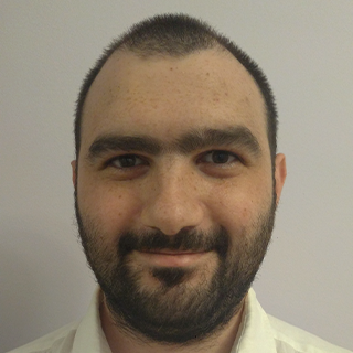 Front-facing portrait of a smiling man with short dark hair, a beard, and a white shirt. The background is plain and light-colored.