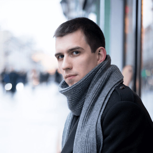 A young man wearing a grey scarf and black coat, looking intently at the camera with a city street blurred in the background.