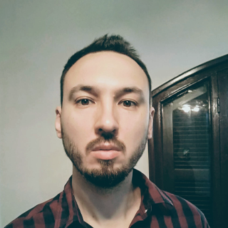 Man with short hair and stubble wearing a plaid shirt, standing indoors with a neutral expression.