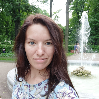 A woman with shoulder-length brown hair smiling at the camera with a fountain in the background and trees surrounding the area, indicating a park setting.