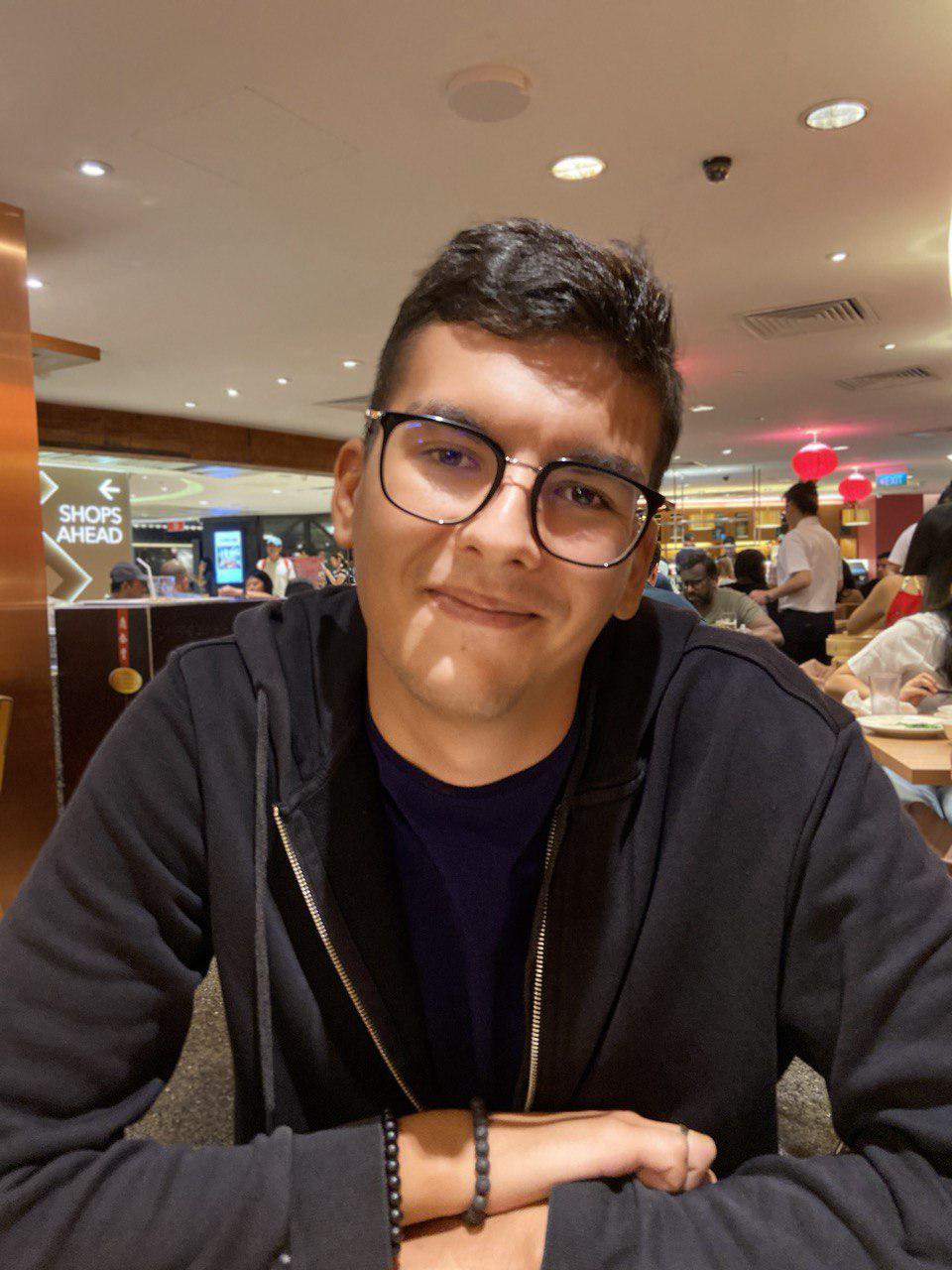 Young man with glasses smiling at the camera, seated in a busy food court with a blurred background. He's wearing a black jacket over a blue shirt, with a black bead bracelet on his wrist.