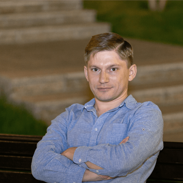 A man in a blue shirt sitting on a bench with arms crossed, smiling slightly at the camera, with soft-focus stairs in the background.