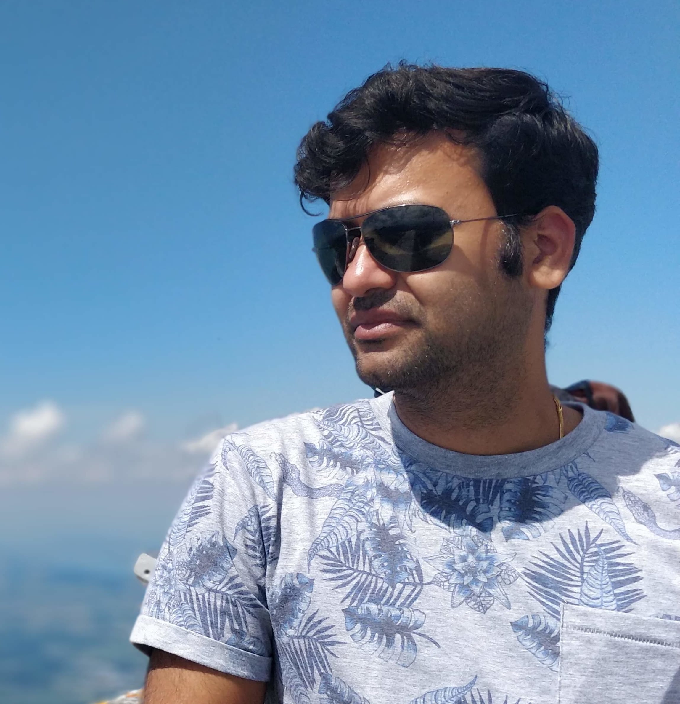 Man with sunglasses looking to the side, wearing a patterned shirt with a clear sky background.