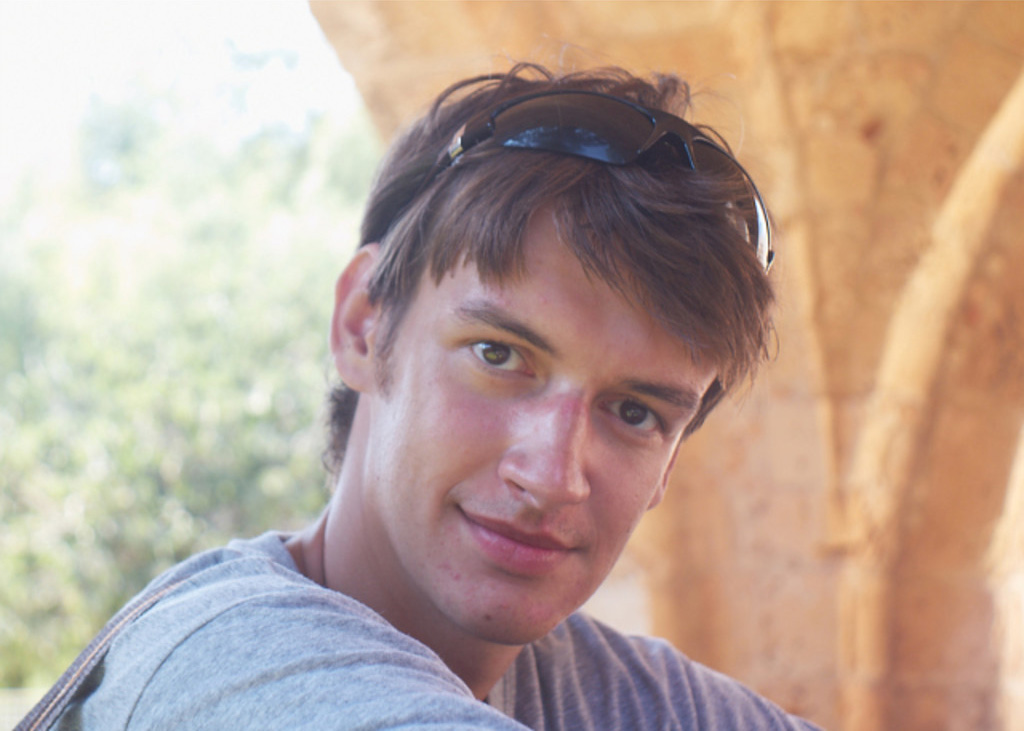 A close-up of a young man with sunglasses on his head, smiling slightly at the camera, with soft-focus natural and architectural background.