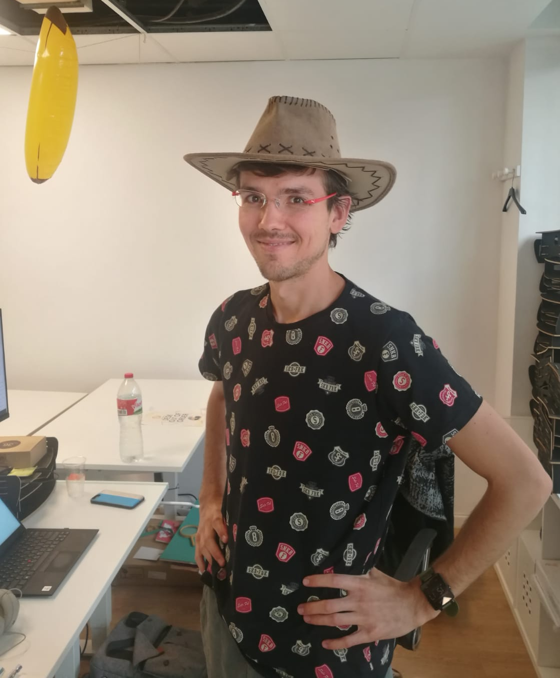 "Man smiling while wearing a cowboy hat and red-framed glasses, standing in an office with a yellow pendant light hanging overhead and laptops on desks in the background."