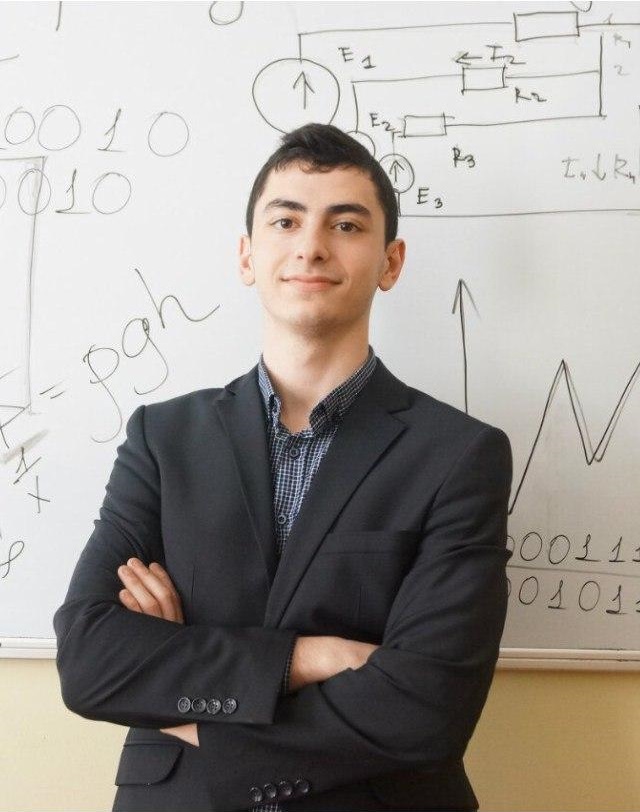 A young man in a business suit standing in front of a whiteboard with mathematical and electrical diagrams.