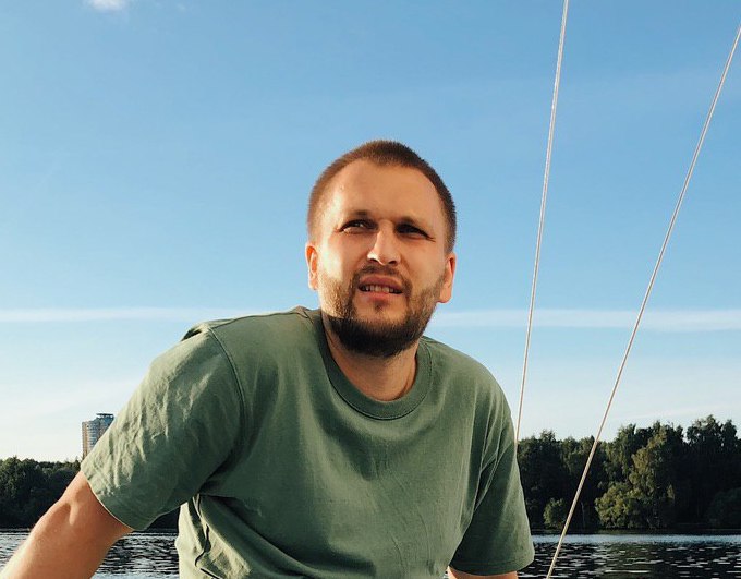 A man in a green t-shirt squinting in the sunlight on a boat with water and trees in the background.