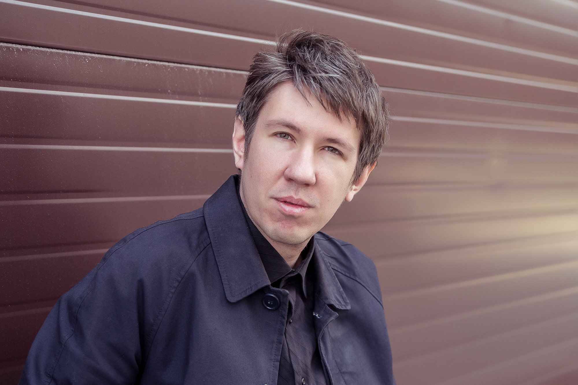 Young man with a contemplative expression, wearing a dark jacket, against a blurred brown corrugated metal background.