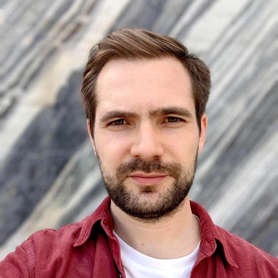 Selfie of a man with a beard and moustache, wearing a red shirt over a white tee, with a blurred background.