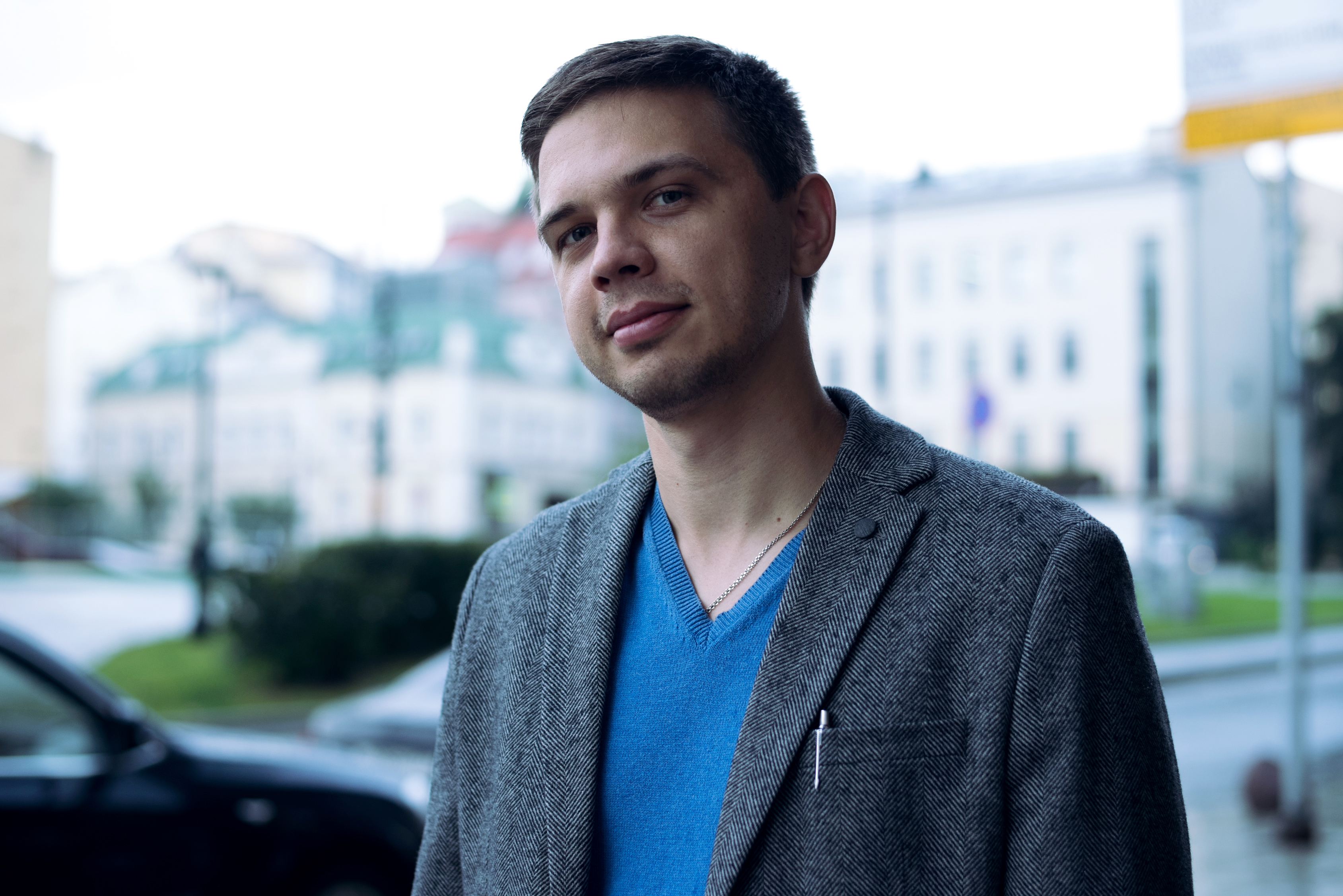 Young man in a grey checked jacket over a blue shirt, standing outdoors with blurred city buildings in the background.