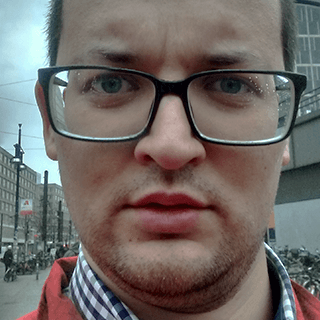 Close-up of a man with glasses looking at the camera with a serious expression, wearing a checkered shirt with a red jacket, with a blurred city street background.