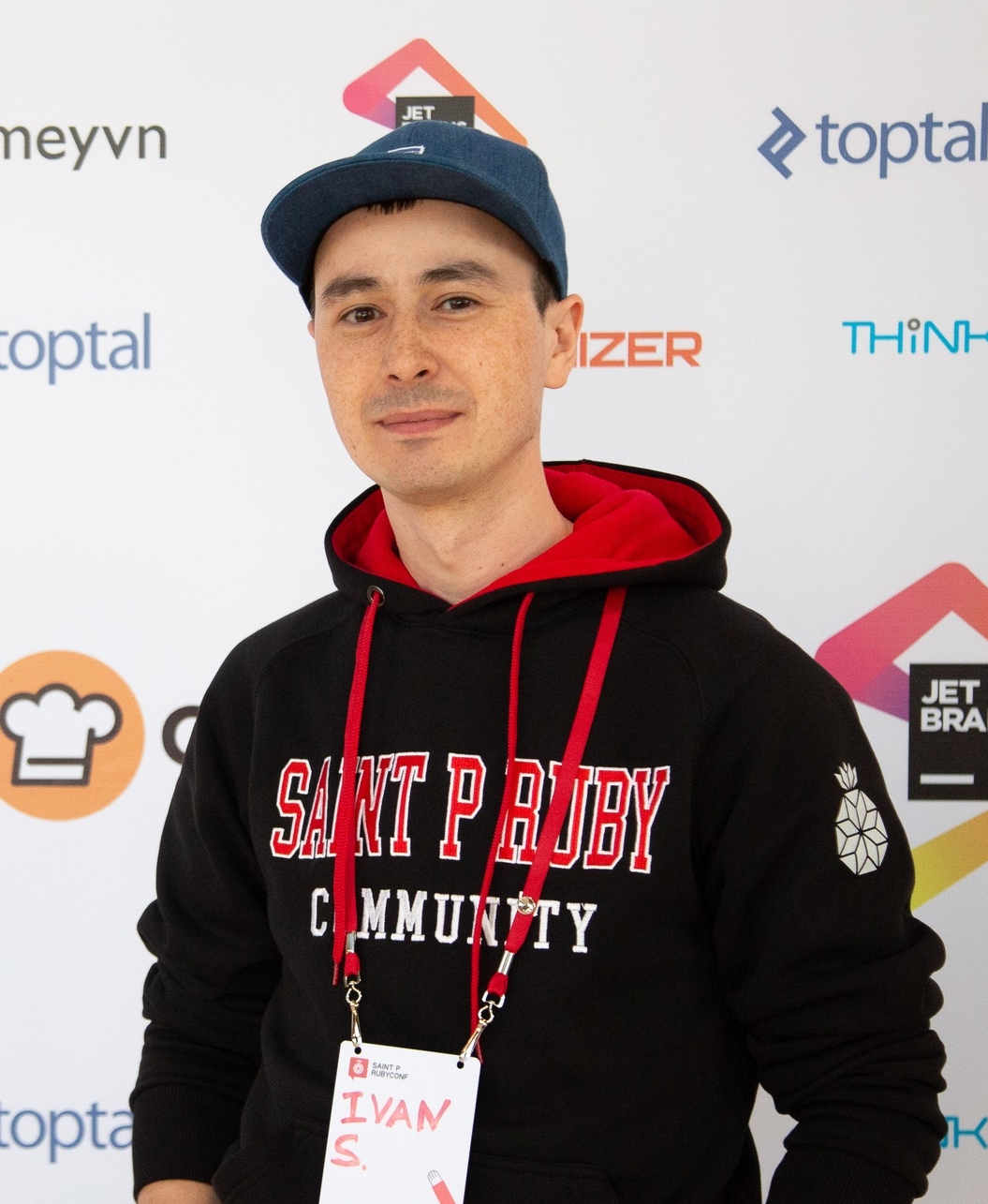 Man in a black hoodie and blue cap standing in front of a backdrop with logos, wearing a badge with the name "Ivan S."