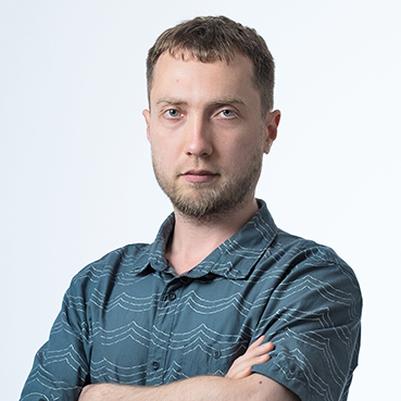 Portrait of a man with short hair and stubble, wearing a patterned shirt, arms crossed, looking directly at the camera with a serious expression, against a white background.