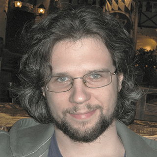 Close-up of a smiling man with medium-length curly hair, glasses, and a goatee, dressed in a casual jacket, in an indoor setting.