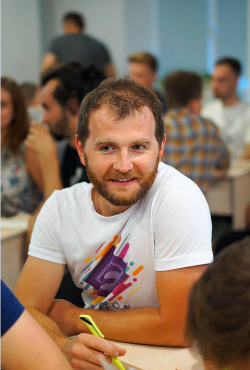 Man in a white T-shirt with colorful logo sitting in a busy room with people in the background. He has a beard, is smiling, and appears to be engaged in a conversation.
