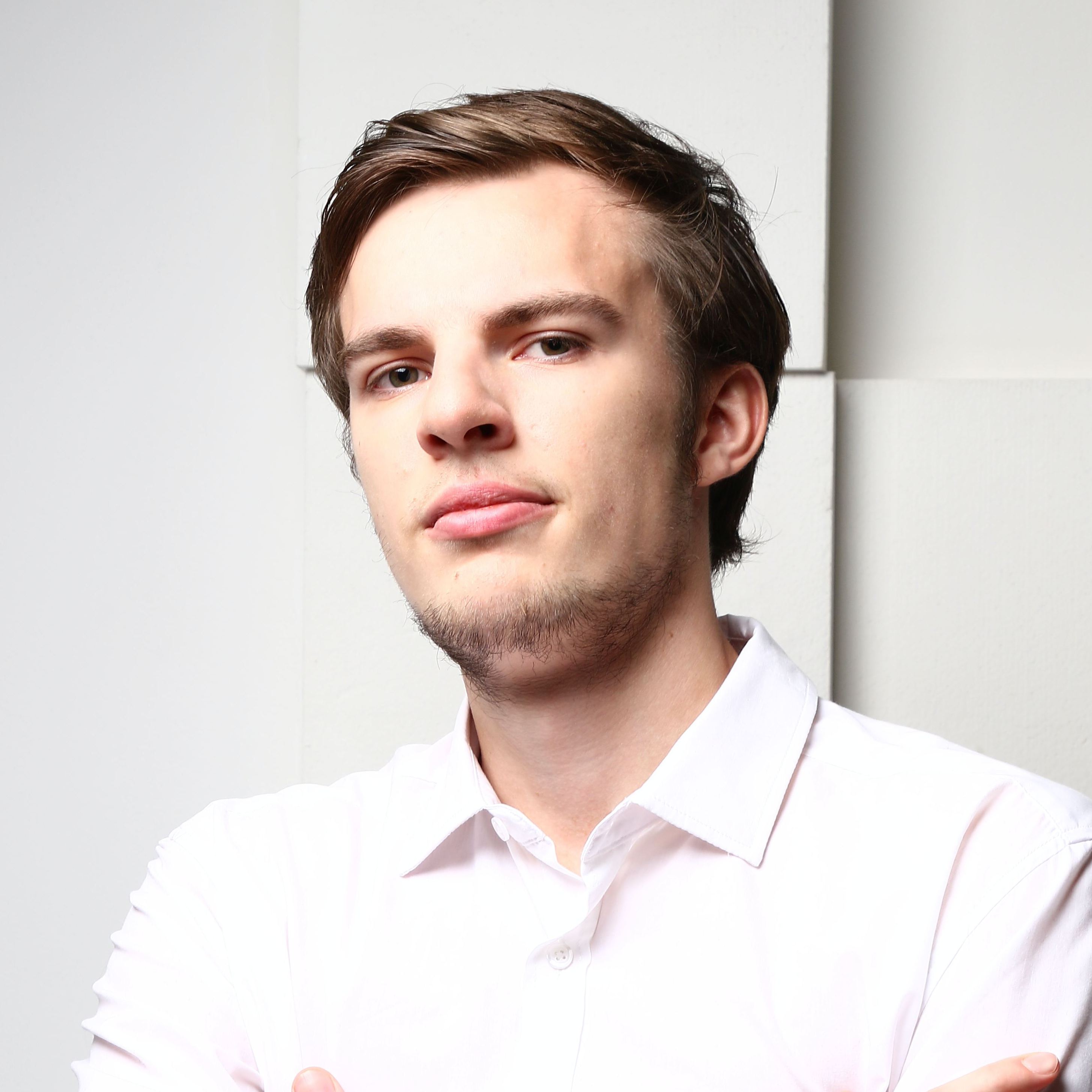 Headshot of a young man with a slight beard wearing a white shirt, standing against a white backdrop, looking at the camera with a serious expression.