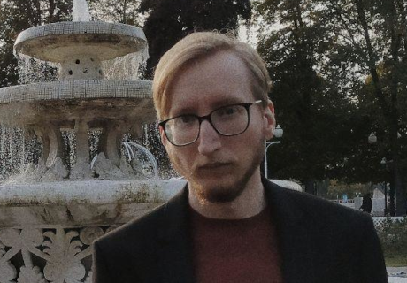 A person with glasses and a beard standing in front of a fountain with water cascading down multiple levels, surrounded by trees in a park setting during twilight.