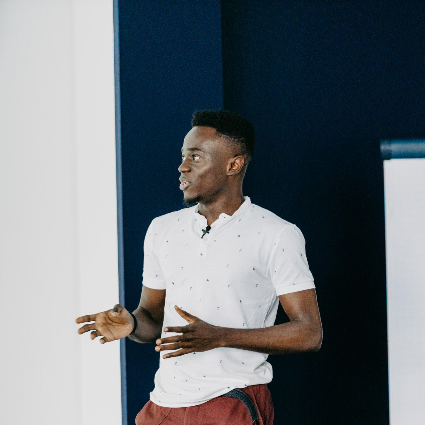 A confident young man presenting or speaking in front of a dark blue and white background, gesturing with his hands.