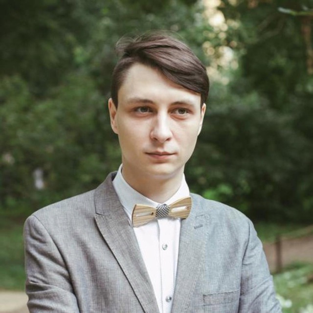 A young man with short hair, wearing a grey suit and bow tie, standing in front of a green, leafy background.