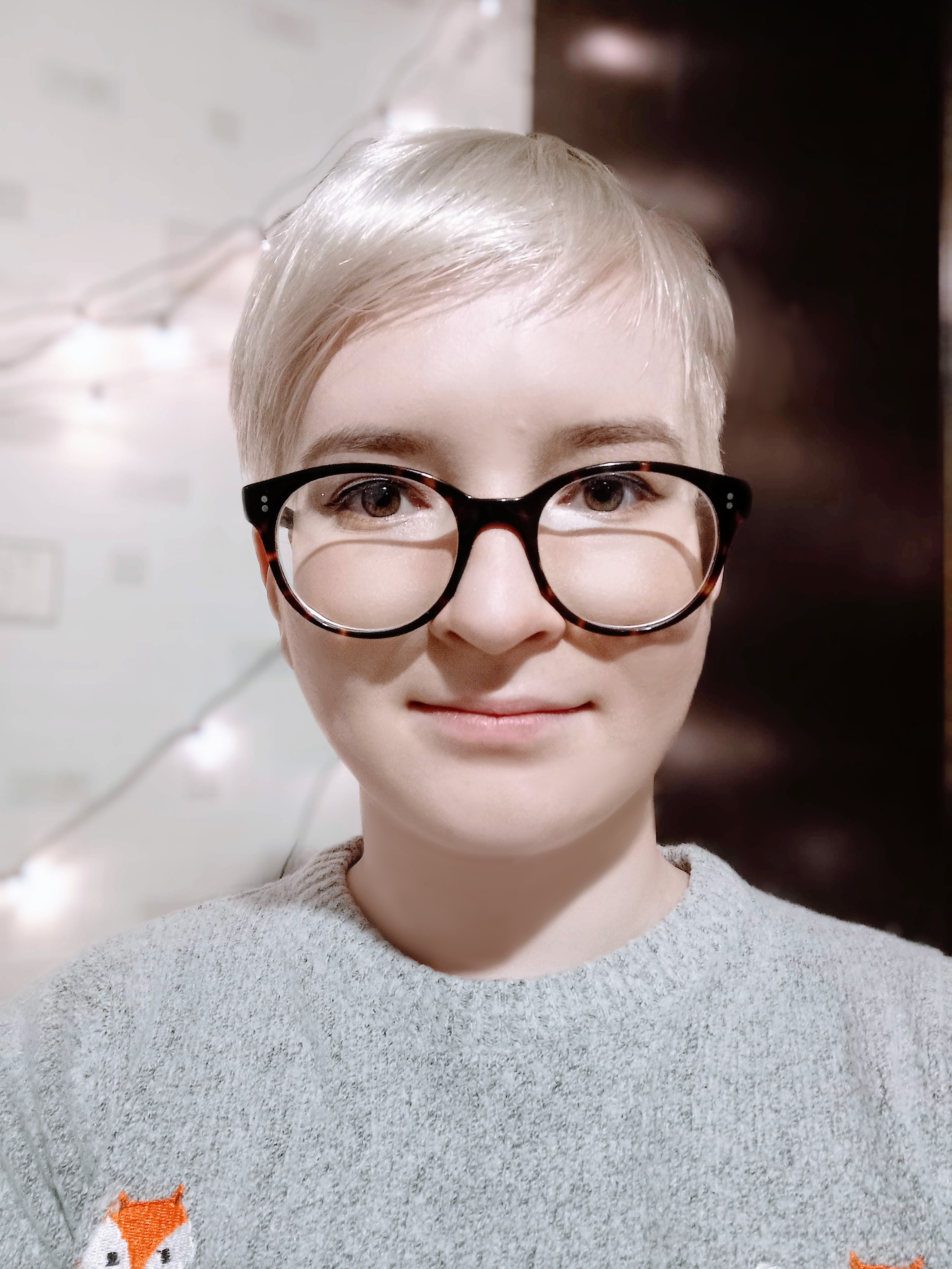 Person with short platinum blonde hair wearing black-rimmed glasses and a grey sweater with a fox design, set against a bokeh light background.