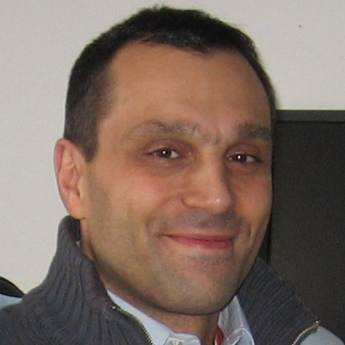 Close-up of a smiling man with short dark hair wearing a grey zip-up sweater and a collared shirt, indoors with a neutral background.