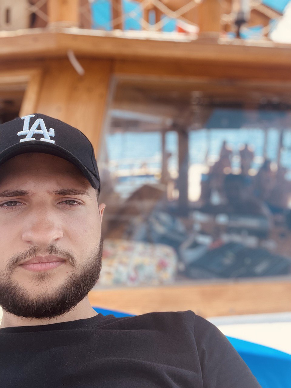 "Man in a black LA cap and t-shirt taking a selfie with a wooden structure and blurry background on a sunny day."