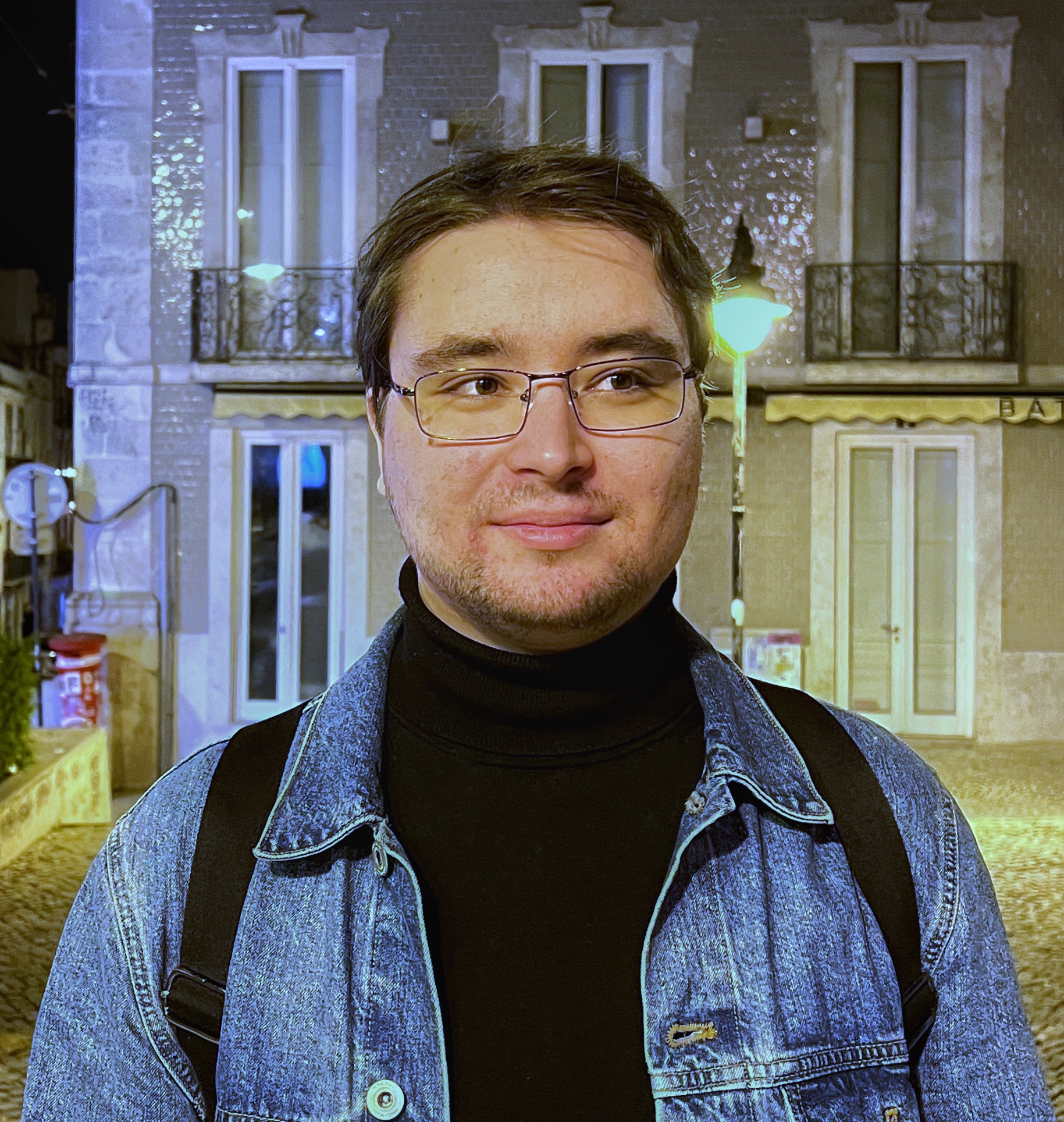 A person with glasses smiling subtly at the camera, wearing a turtleneck and a denim jacket, with urban night-time scenery in the background.